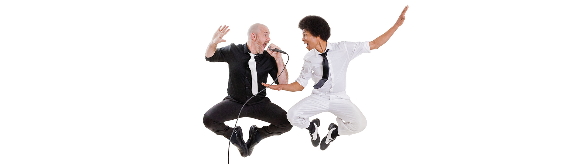Two men in a mid-air jump against a white background, with the man on the left wearing a black shirt and pants with a white tie and the man on the right wearing a white shirt and pants with a black tie. The man in black is singing into a microphone while the man in white has his arms outstretched.