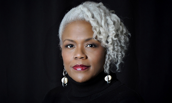 Portrait of Marietta Simpson with curly silver hair, wearing a black turtleneck and distinctive silver earrings, against a dark background.