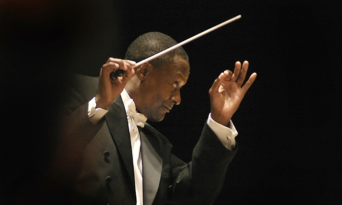 Conductor Thomas Wilkins in formal attire gesturing passionately with a baton, leading an orchestra performance against a dark backdrop.