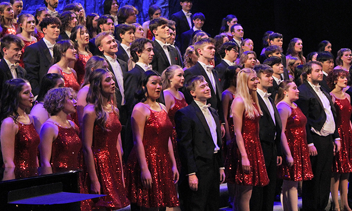 A large choir performs on stage, wearing sparkly red dresses and formal black attire, creating a vibrant visual display.