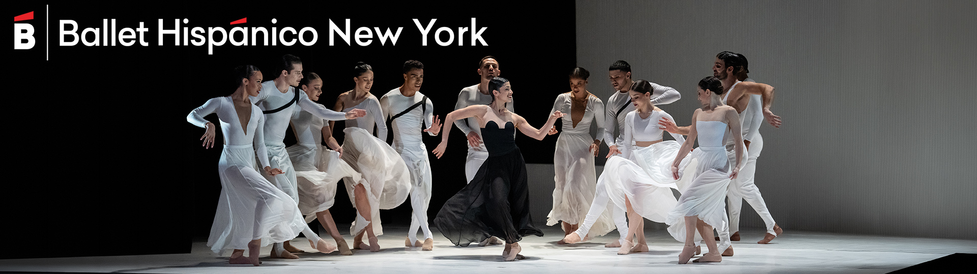 Ballet Hispánico dancers performing onstage in flowing white costumes, surrounding a central dancer in black, with the company logo above.